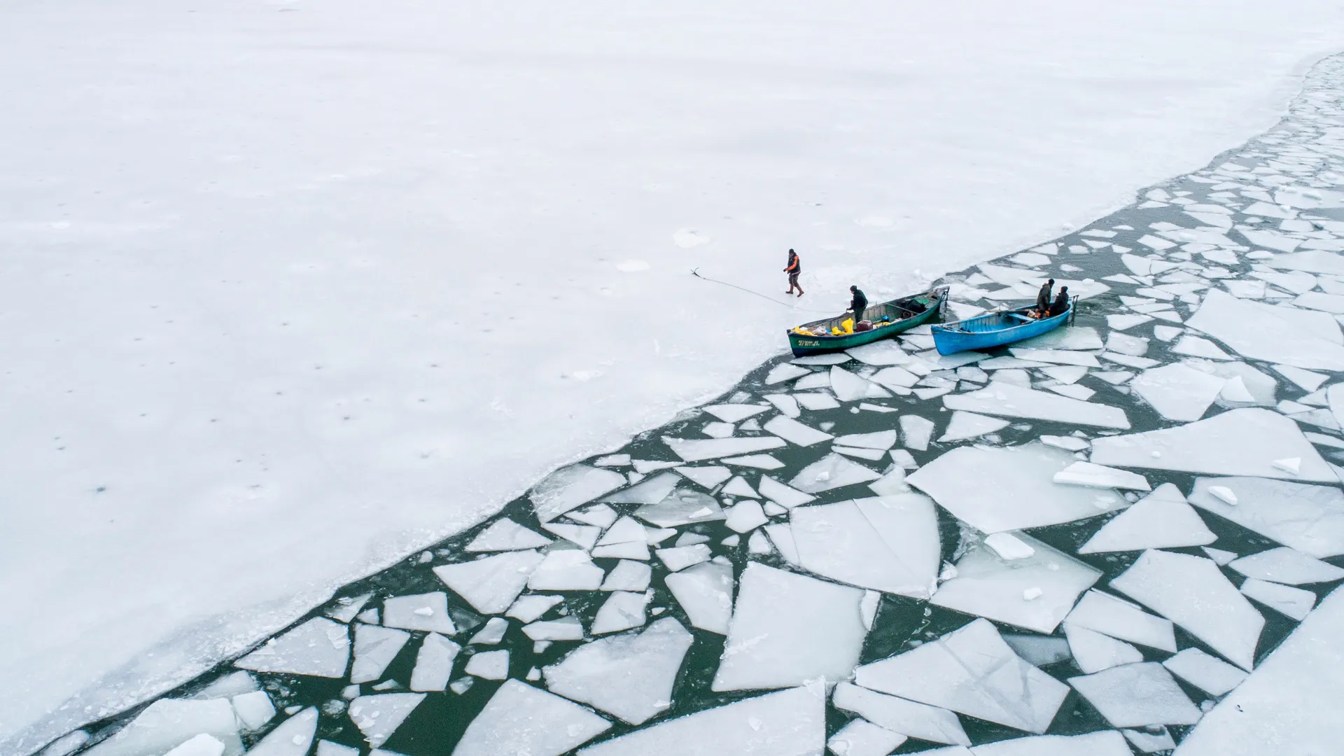 Aerial view of two small fishing boats at the edge of a frozen sea, where solid ice breaks into geometric floes over dark water